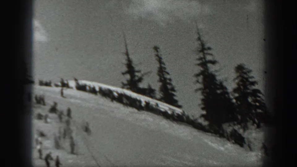 Distant skiers occupy a snowy Washington slope below tall evergreens and a dark ridge line in a grainy 8mm frame.
