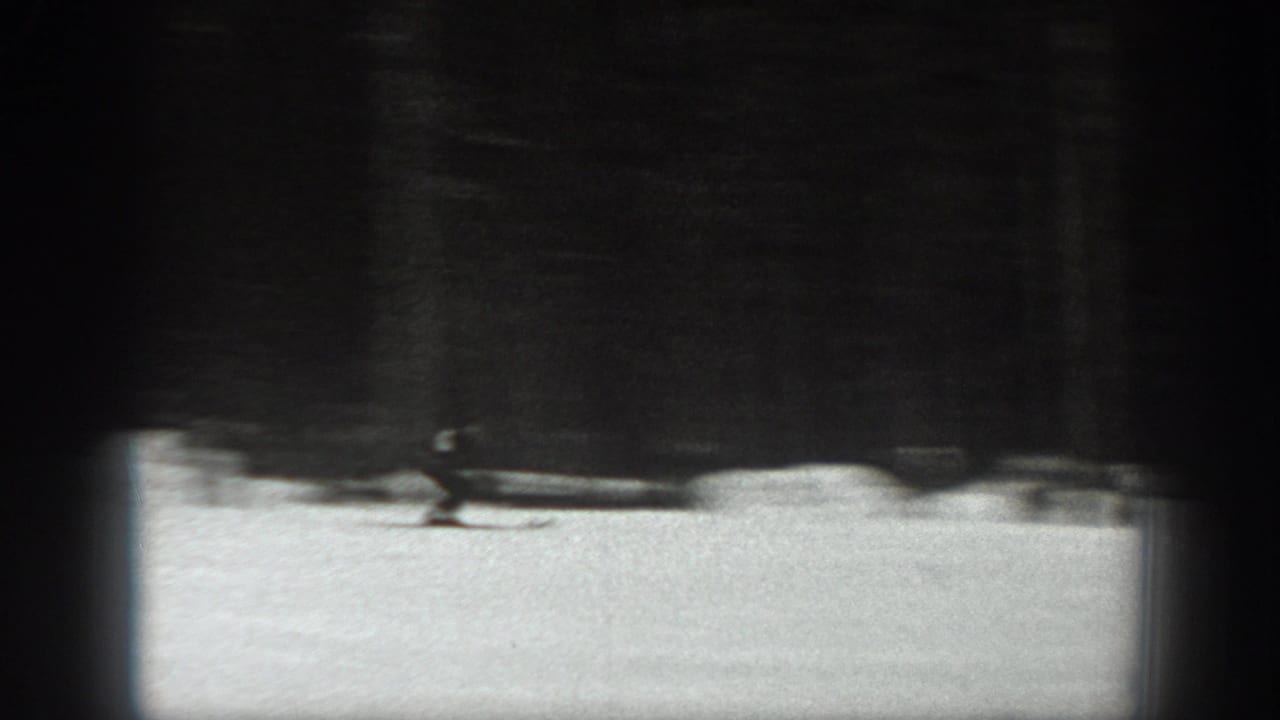 A small skier figure crosses a bright snowy area below dark trees or a structure.