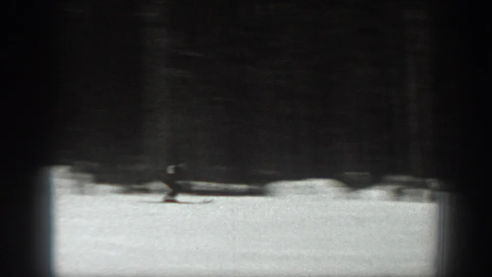 A distant skier appears on snow beneath a dark tree line or slope structure in grainy 8mm footage.