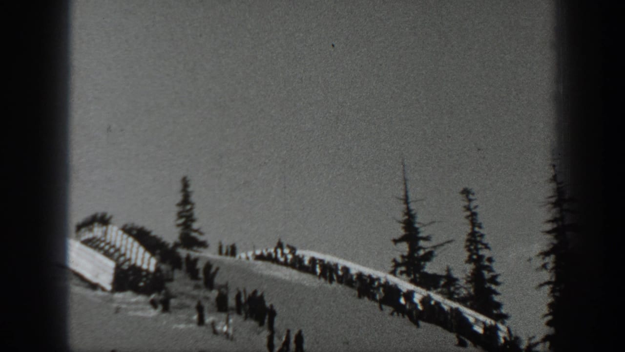 Snowy ridge with spectators, evergreen trees, and a ski slope seen through dark film edges.