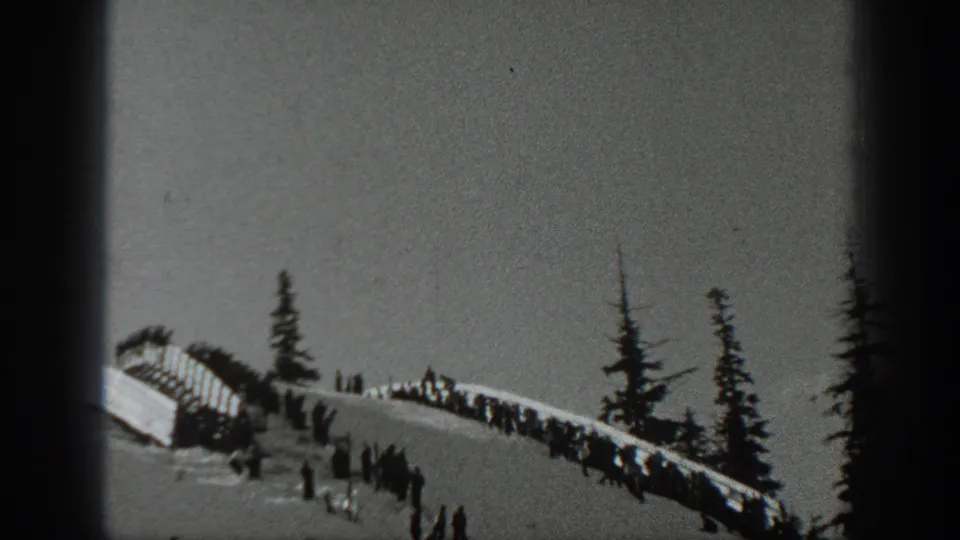 Washington 1940 home-movie frame showing a snowy ridge, distant onlookers, evergreen trees, and a ski slope in black and white.