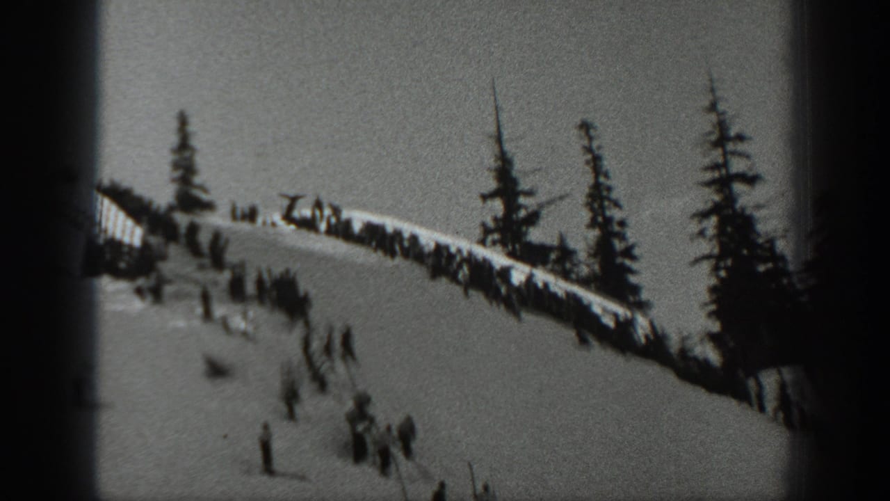 Snow ridge with clustered spectators and tall evergreens beside a descending ski slope.