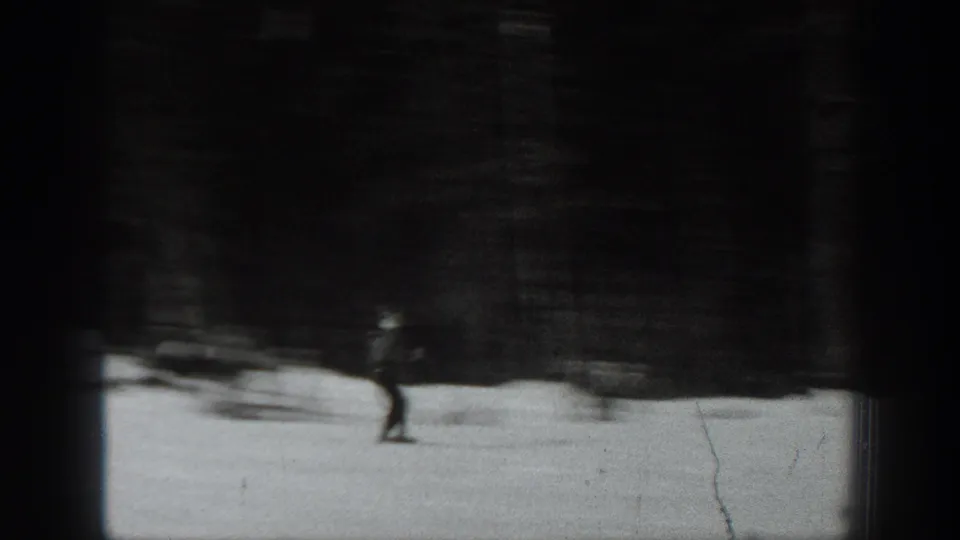 Black-and-white 8mm frame of a distant skier gliding across snow in Washington, 1940, with a dark background behind.