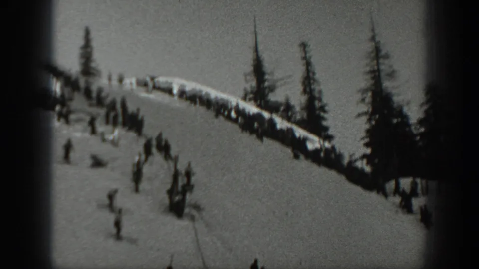 Washington 1940 frame with grouped spectators beside a snowy ski route, evergreen trees, and distant skiers in soft focus.
