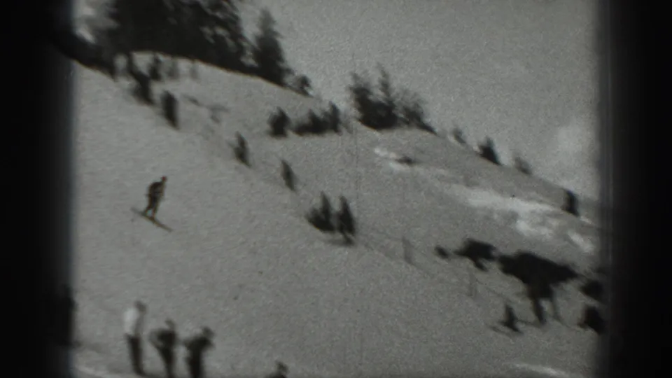 A distant skier travels across a Washington ski slope in 1940, with small spectator figures, snowbanks, and trees nearby.