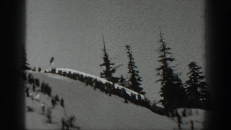 Black-and-white 8mm frame of a Washington snowy ridge with onlookers above riders moving downhill.