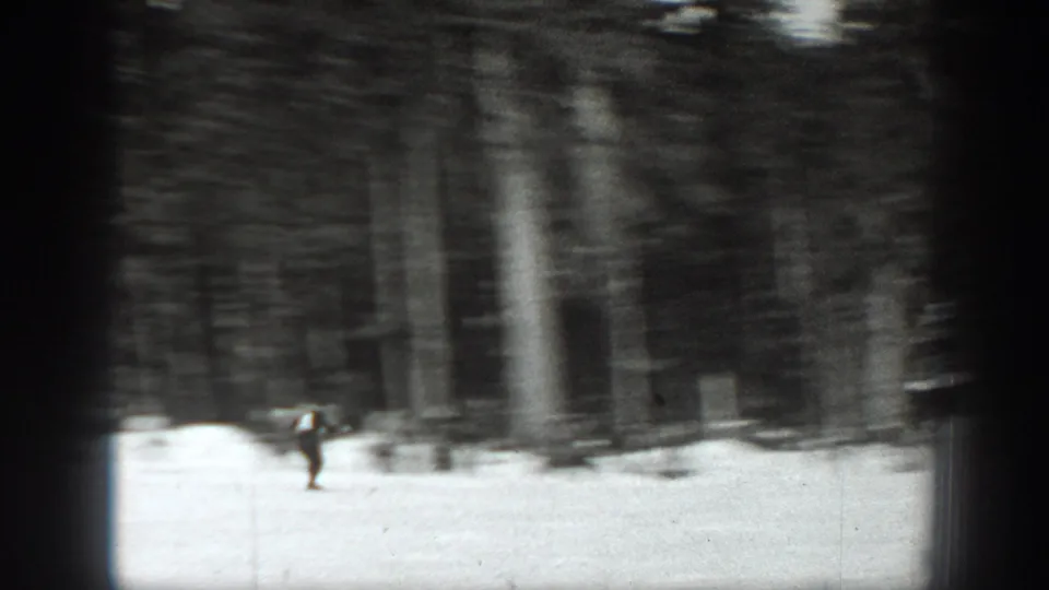 A distant skier moves across a snowy Washington slope in black-and-white 8mm footage, with dense evergreen trees behind him.