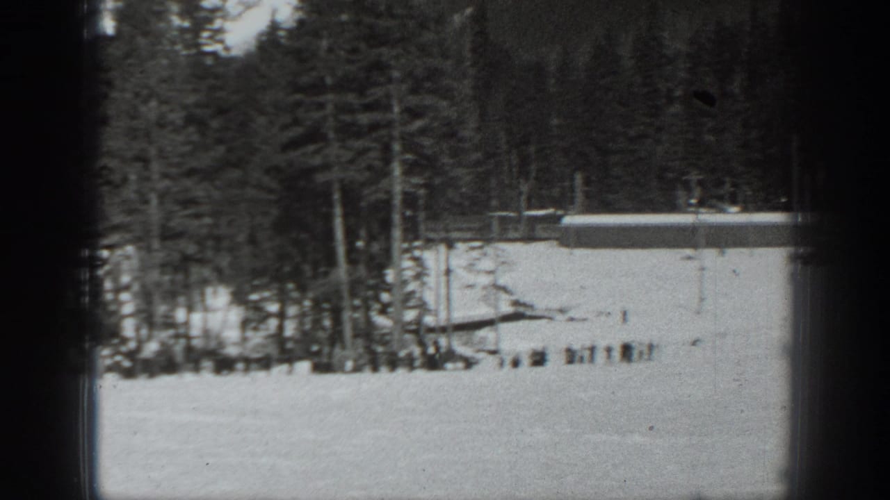 Snowy open slope with evergreen trees and a low lodge structure in the background.