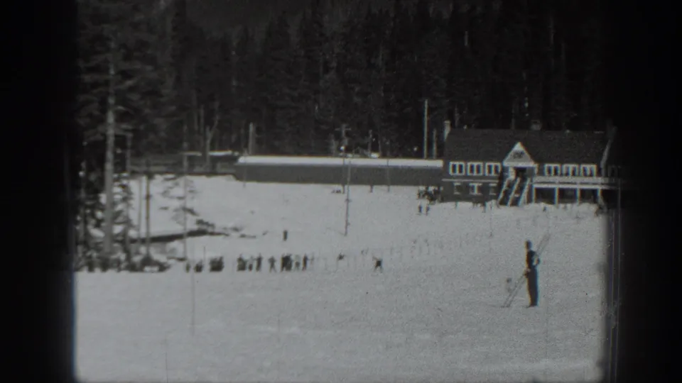A skier stands on a snowy Washington slope, with lodge buildings, poles, evergreen trees, and small distant groups in the background.
