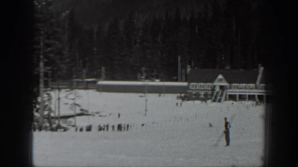 The skier remains on the snowy Washington slope below lodge buildings, with evergreen woods and distant figures visible in the 8mm frame.