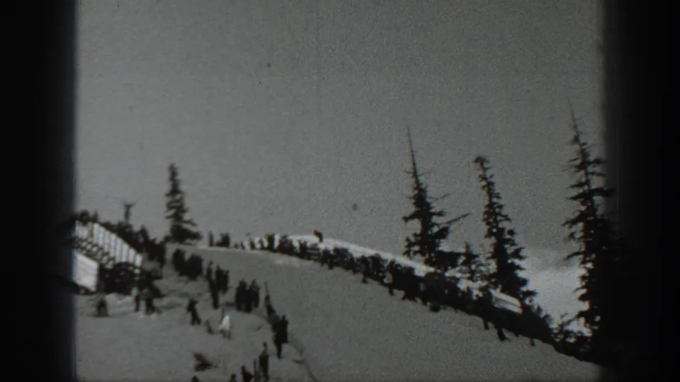 1940 Washington frame showing a snowy ski-event ridge with spectators, evergreen trees, and a distant skier or jumper near the lower edge.
