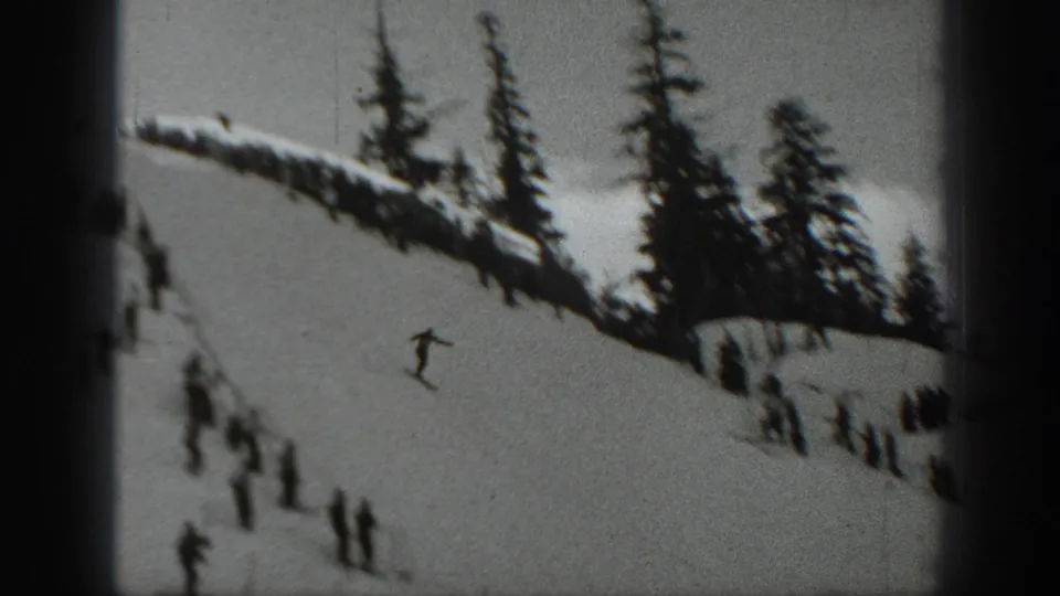 A lone skier moves downhill on a snowy Washington mountain slope, with dark evergreens and distant figures nearby.