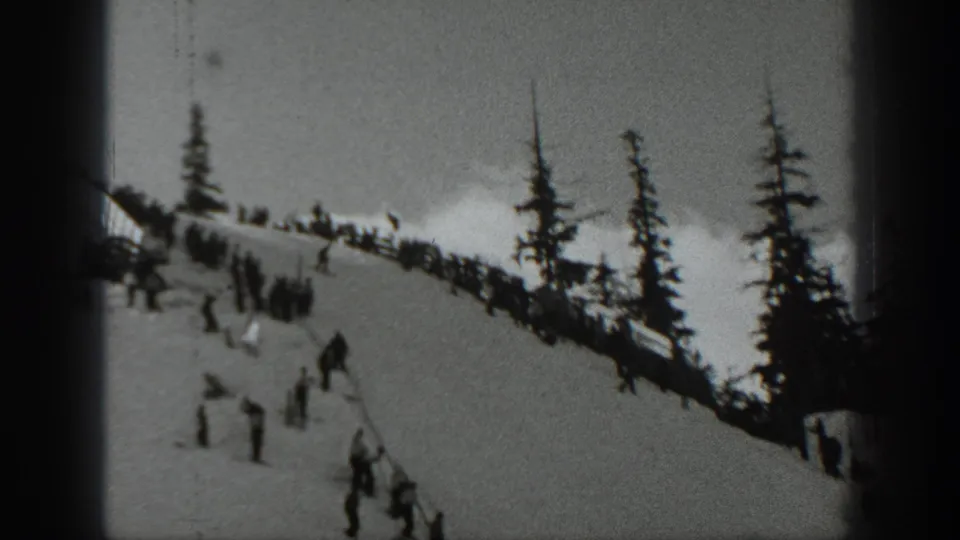 Skiers and spectators cluster on a Washington snow slope, with tall evergreens and soft black-and-white 8mm texture.