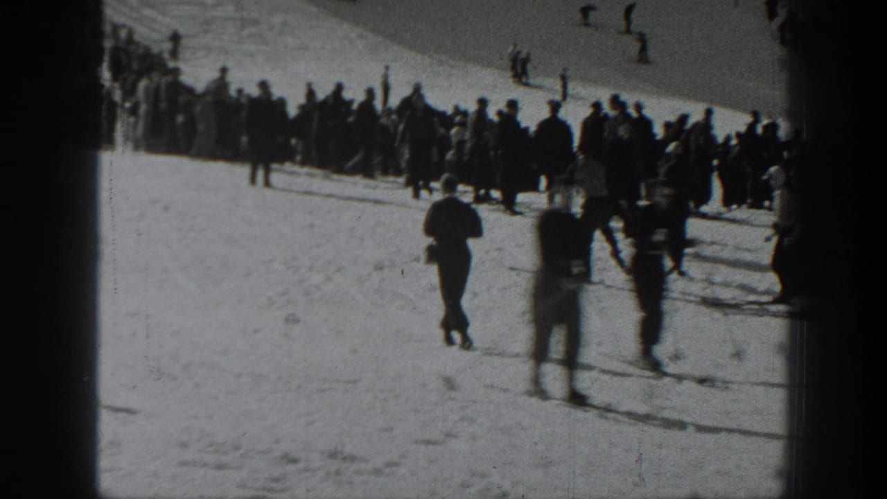 Crowd stands beside a snowy ski slope as skiers pass in the foreground.