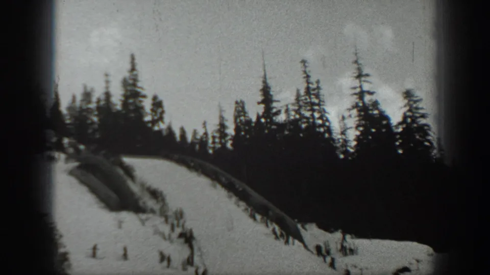 Soft black-and-white 8mm frame of a snowy Washington slope bordered by evergreens, with distant skiers below.