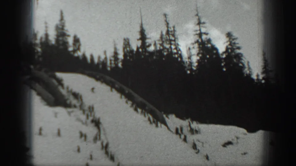 1940 Washington 8mm frame showing a steep snow-covered slope, dark pines along the ridge, and small skiers moving downhill.