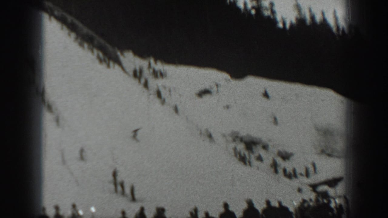 Wide snowy slope with distant skiers and a dark forested ridge at the top.