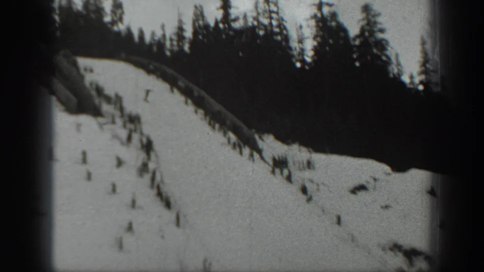 Home-movie frame from 1940 Washington showing skiers scattered across a steep snow slope under a pine-lined ridge.