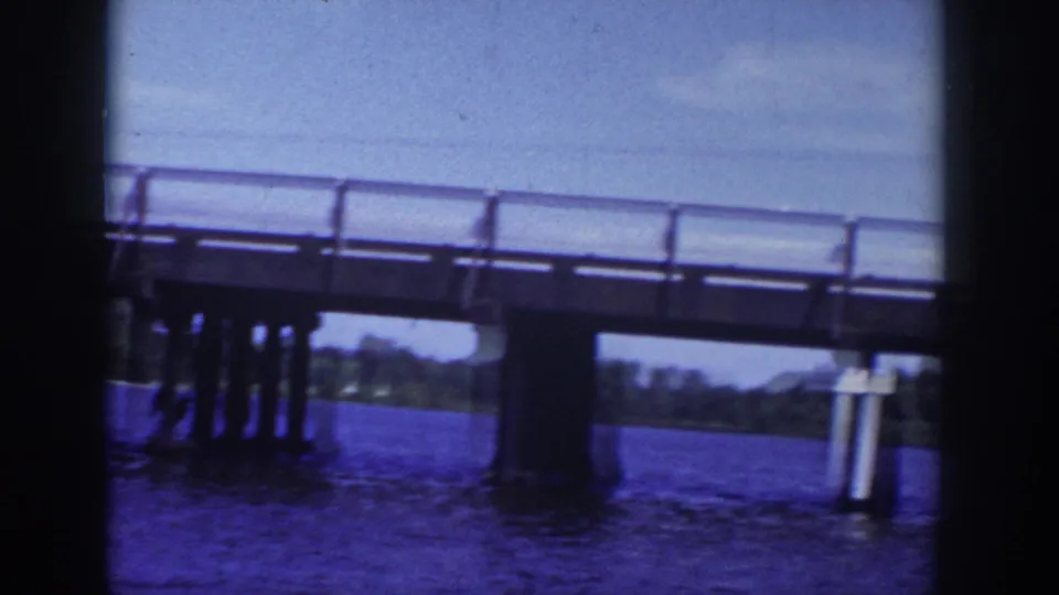 Blue-tinted 8mm frame of a Wisconsin bridge span over water, with structural supports and a distant shore visible.
