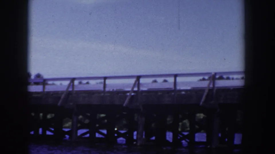 End frame of the Wisconsin bridge scene, showing railing, support structure, water, and distant shoreline in soft 8mm color.