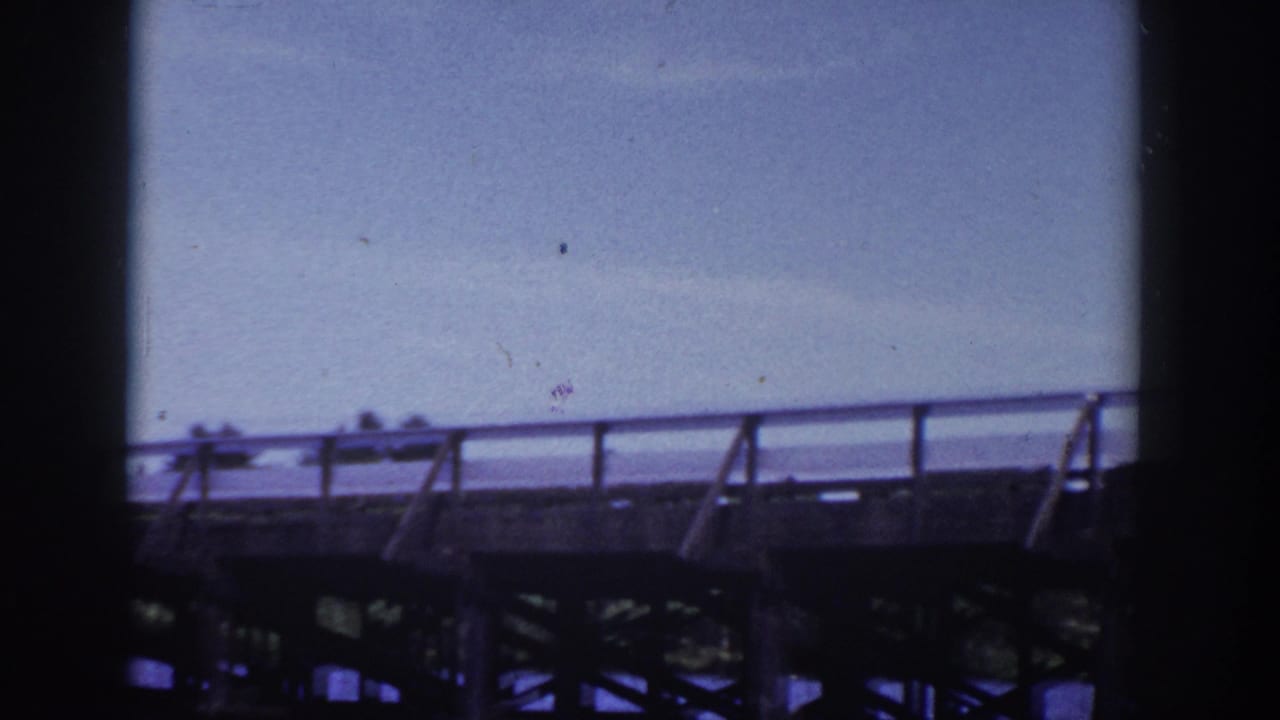 Wooden bridge or dock crossing water beneath a pale sky, seen from a moving boat.
