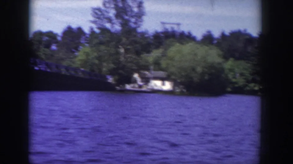 1953 Wisconsin lake shoreline with dense trees, rippled water, and a small lakeside building seen from the boat.