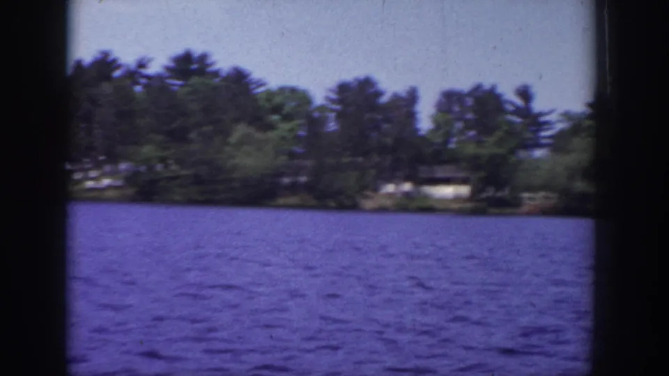 Soft 1953 Wisconsin home-movie frame of rippled lake water, wooded shore, and low lakeside buildings.