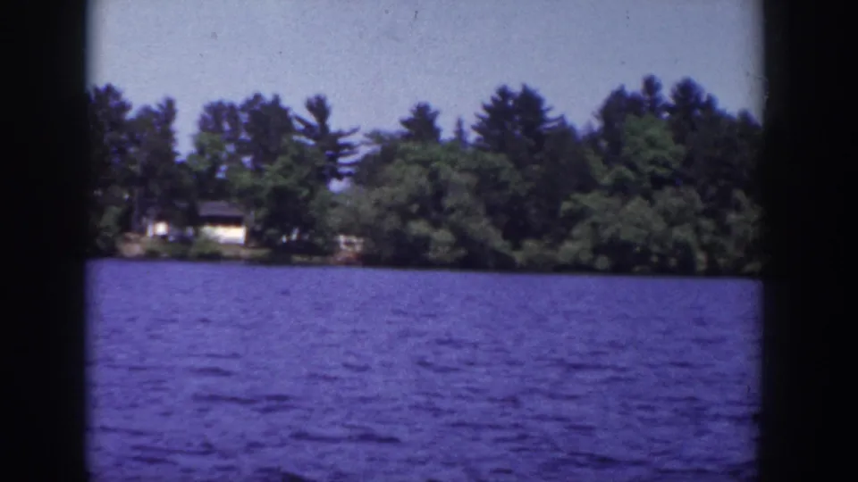 1953 Wisconsin 8mm frame showing rippled lake water, dense trees, and a distant lakeside house across the shore.