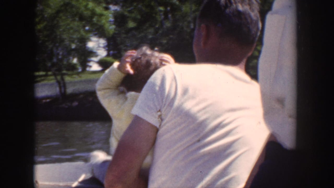1953 Wisconsin 8mm frame showing a man in a light shirt with a baby beside him as the boat passes the wooded shore.