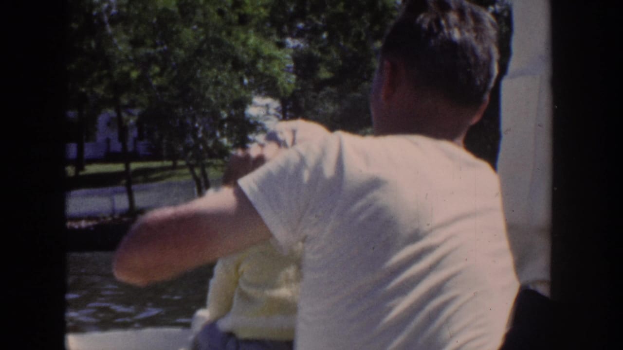 1953 Wisconsin home-movie frame with a man viewed from behind on a boat as a baby sits close beside him near the water.