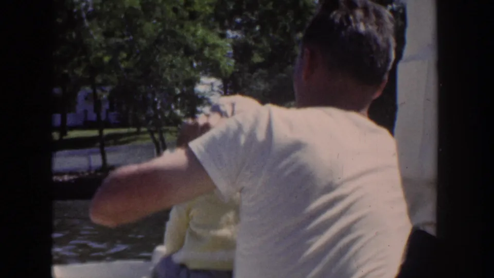 1953 Wisconsin home-movie frame with a man viewed from behind on a boat as a baby sits close beside him near the water.