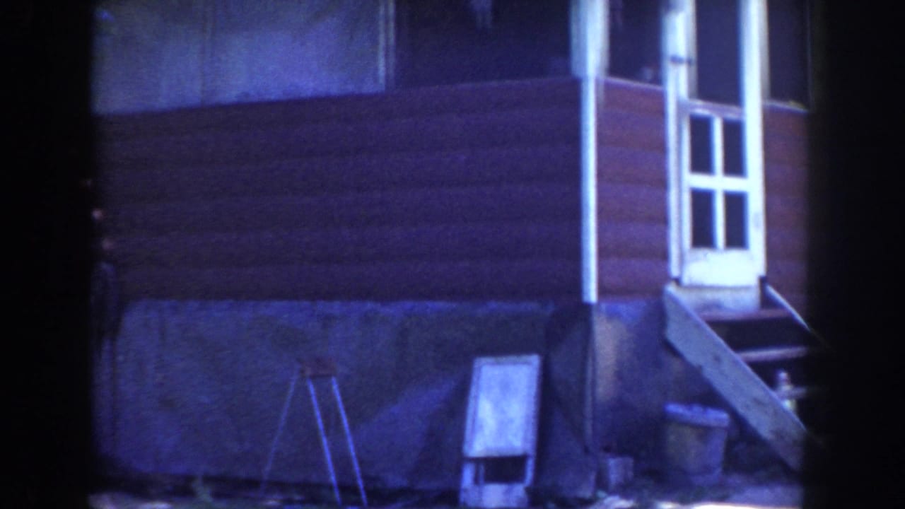 1953 Wisconsin 8mm frame showing a red rural house exterior with white trim, porch steps, basement opening, and yard tools near the wall.