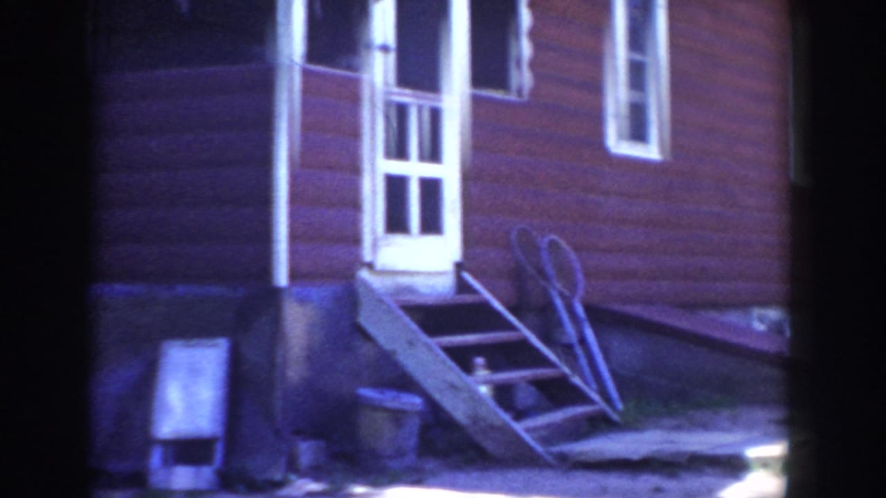 Red house exterior with white door and window, porch steps, and a leaning ladder or yard tool on the grass.