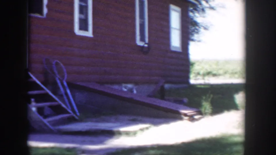 1953 Wisconsin 8mm frame showing the side of a red farmhouse, white windows, porch steps, lawn, and sunlit yard beyond the wall.