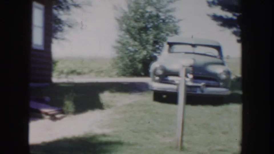 1953 Wisconsin home-movie frame with the red farmhouse edge, grassy lawn, shrubs, and a rounded mid-century automobile parked outside.
