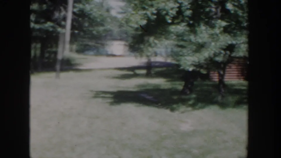 1953 Wisconsin 8mm frame showing a shaded yard, leafy trees, and a log-sided cabin-like building partly obscured.