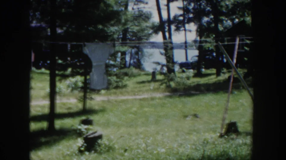 1953 Wisconsin 8mm frame showing a lakeside yard with a garden trellis, white fabric on a clothesline, trees, and water behind.