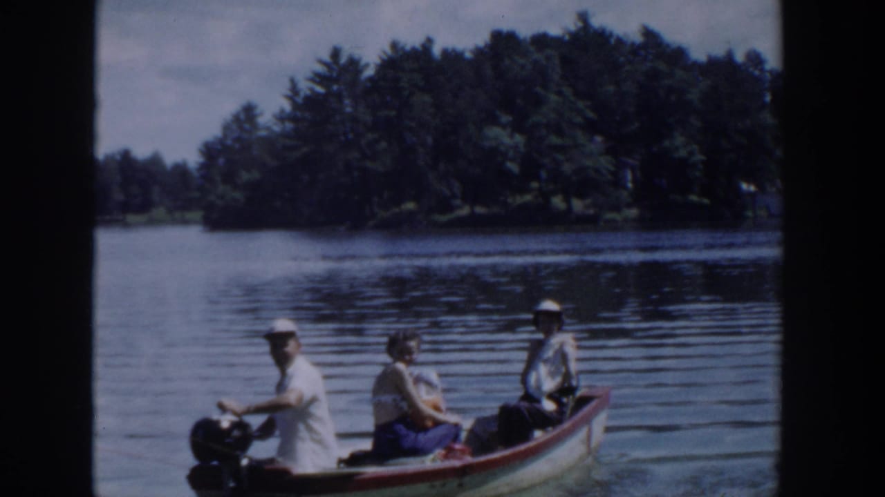 1953 Wisconsin 8mm frame showing three adults in a small motorboat crossing a calm, tree-lined lake.