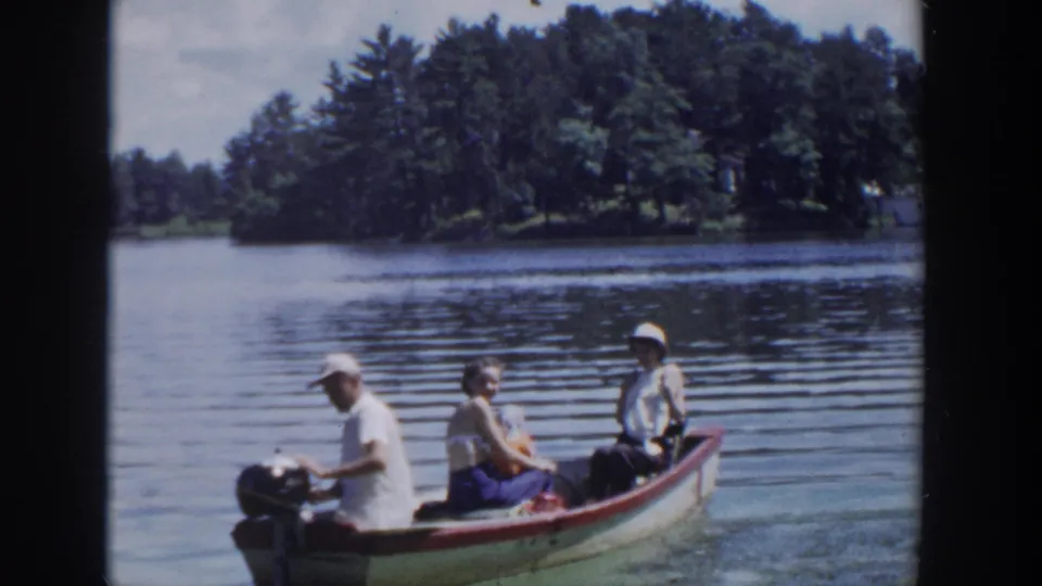 1953 Wisconsin 8mm footage frame of three adults boating on a sunny lake with a tree-covered shoreline behind them.