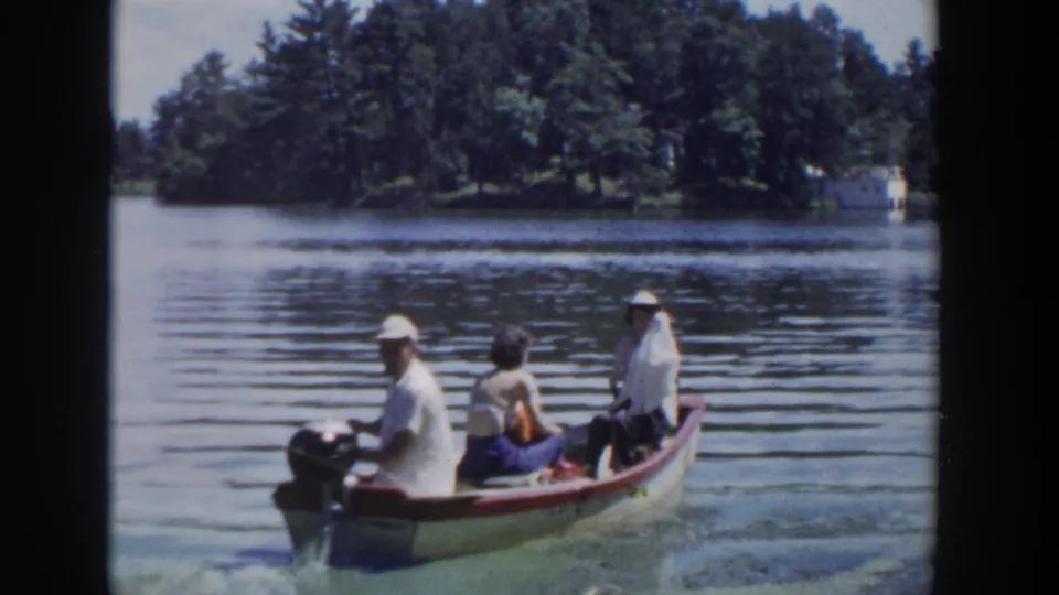 1953 Wisconsin home-movie frame showing three adults in a small motorboat moving across a calm lake near trees.