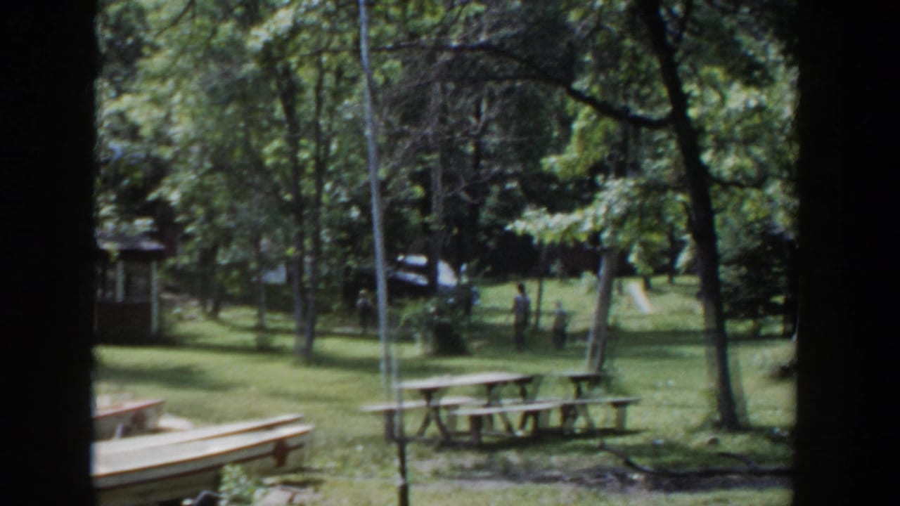 1953 Wisconsin home-movie frame showing a shaded park lawn with picnic tables, canoes near the grass, and a small shelter among trees.