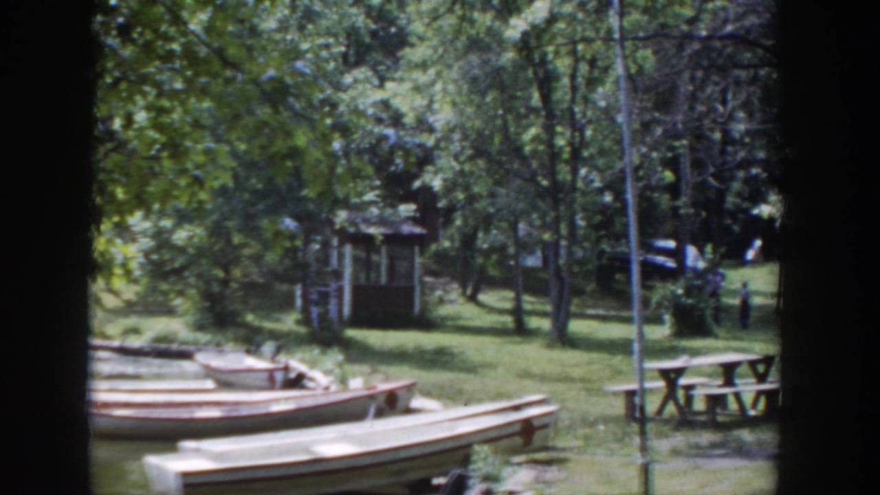 Wooden canoes rest near a grassy lakeside path with trees and a small shelter behind them.