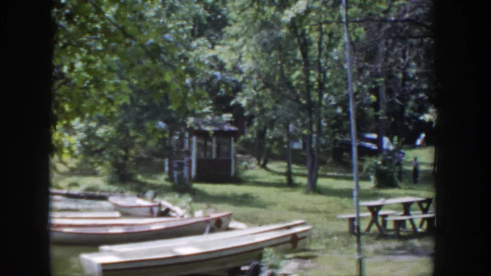 1953 Wisconsin park footage frame with several wooden canoes near the water edge, trees, picnic tables, and a small shelter in the background.