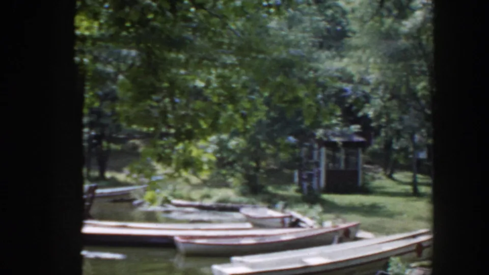 1953 Wisconsin home-movie still showing canoes along a shaded waterway, with a park shelter and picnic area partly obscured by foliage.