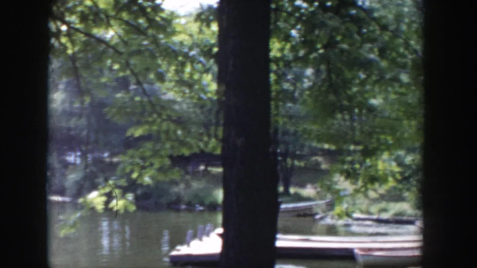1953 Wisconsin lakeside park frame with calm water, docked canoes, dense trees, and a dark foreground trunk framing the view.