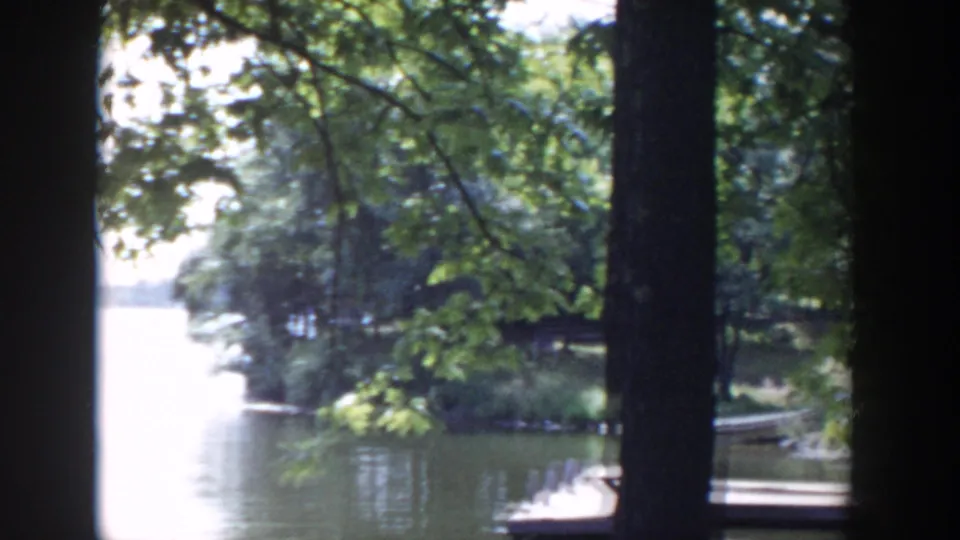 1953 Wisconsin 8mm frame looking through lakeside trees toward calm water, a wooded shoreline, and a small dock.