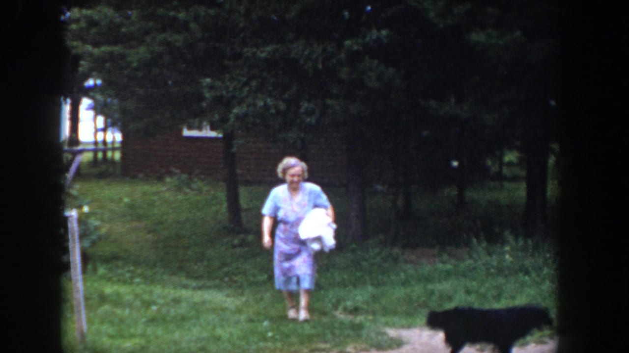 In a 1953 Wisconsin home-movie frame, a woman in a light dress carries a white bundle across a green yard while a black dog moves nearby.