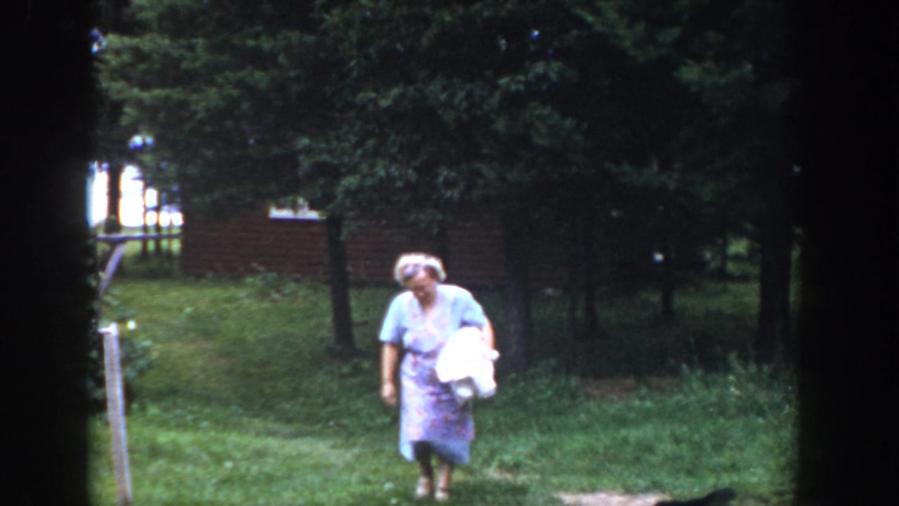 Woman in a pale dress walks through grass holding a white bundle near trees and a brick wall.