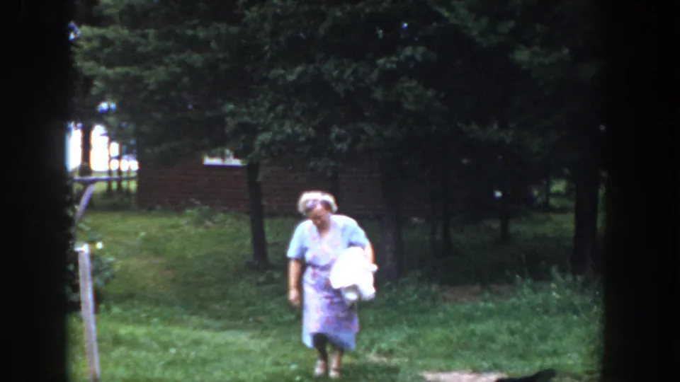 A woman in a pale dress moves through a green Wisconsin yard in 1953, holding a white bundle with trees and brickwork behind her.