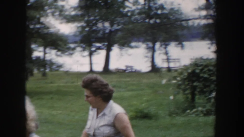 A woman with glasses appears in profile in a 1953 Wisconsin yard, with grass, trees, and a body of water visible beyond her.